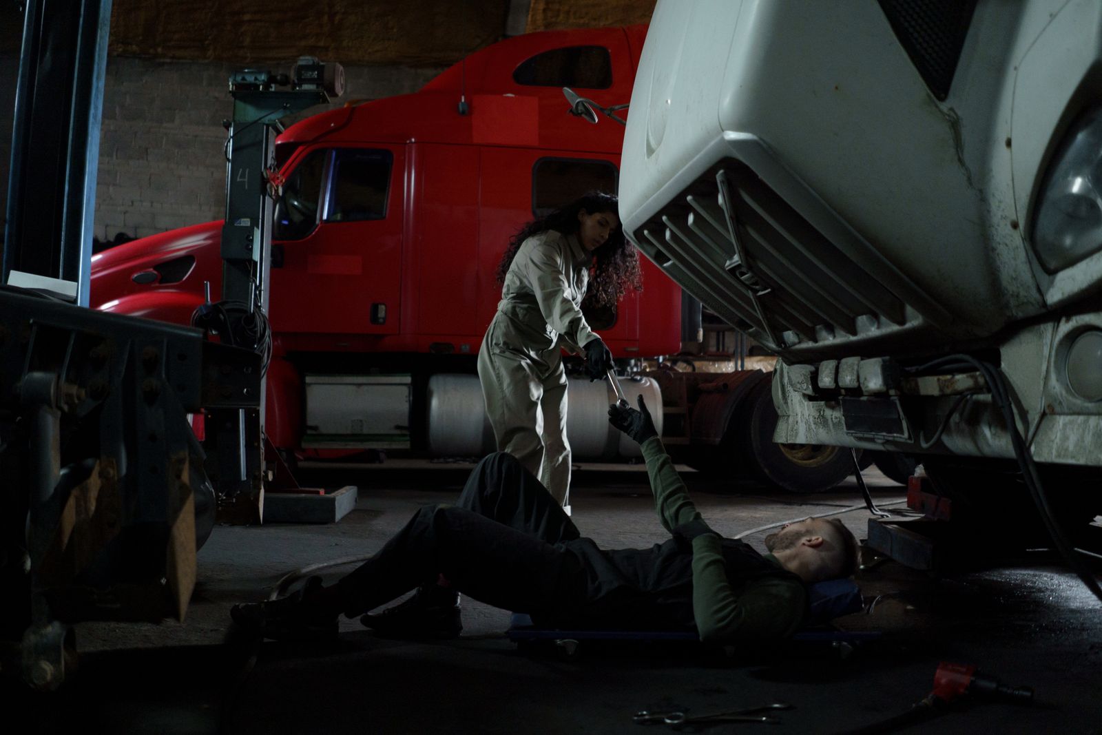 Two mechanics repairing a heavy-duty semi truck in a shop, one underneath the truck and one handing over a tool